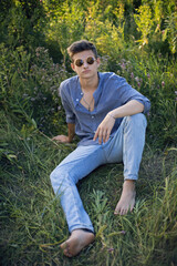 Naklejka na ściany i meble sexy teen guy in sunglasses is sitting on the grass in nature in a shirt and jeans