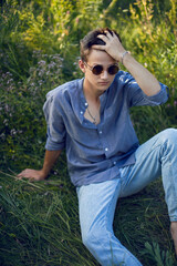 sexy teen guy in sunglasses is sitting on the grass in nature in a shirt and jeans