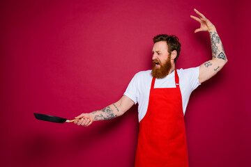 angry chef with beard and red apron cooks with pan - obrazy, fototapety, plakaty
