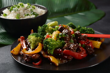 Asian teriyaki beef with red and yellow bell peppers, broccoli and sesame seeds on a plate on the table. Spicy teriyaki beef stir fry with vegetables and rice  on a dark background