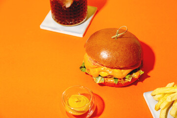 Big burger with french fries and sauce on bright orange and yellow background. American Fast food cuisine in retro style. Copy space