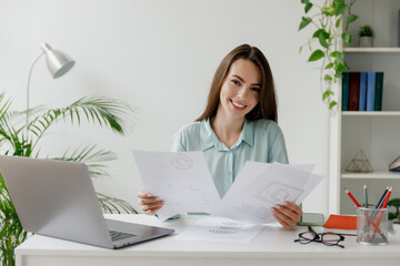 Smiling happy young successful employee business woman in blue shirt hold paper account documents sit work at workplace white desk with laptop pc computer at office indoors Achievement career concept