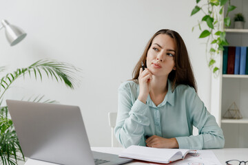 Young minded serious thoughtful employee business woman in blue shirt writes in notebook sit work at workplace white desk with laptop pc computer at modern office indoors Achievement career concept.