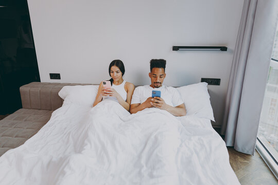 Top View Full Size Body Length Young Couple Two Family Man Woman In Casual White Clothes Lying In Bed Hold Use Mobile Cell Phone Rest Relax Spend Time Together In Bedroom Lounge Home Own Room House