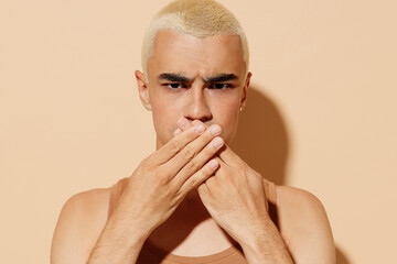 Young shocked frowning secret trendy blond latin gay man with make up in beige tank shirt cover...