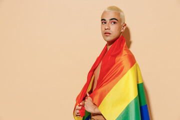 Side view young serious minded blond latin gay man with make up in beige tank shirt wrapped in rainbow flag looking aside isolated on plain light ocher background studio People lgbt lifestyle concept.