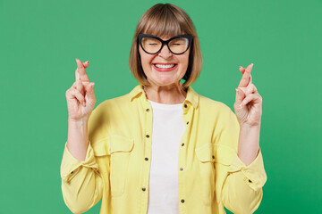 Elderly smiling happy woman 50s in glasses yellow shirt waiting for special moment, keeping fingers...