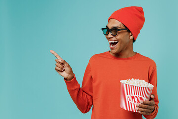 Young african man in 3d glasses red clothes watch movie film hold bucket of popcorn point finger aside workspace isolated on light blue background studio. People emotions in cinema lifestyle concept