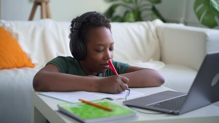 Schoolboy writing and sitting at table with laptop during online learning in home room spbd. Close-up view of African boy writes in notebook and looks at computer screen, speaks with smile and sits at - Powered by Adobe