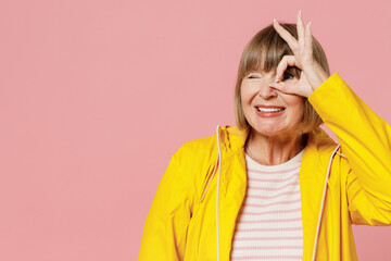 Mature elderly woman 55 years old wears yellow waterproof raincoat show hide eye with okay ok gesture wink isolated on plain pastel light pink background studio portrait. Outdoors wet weather concept.