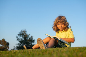 Fototapeta premium Clever child boy reading book laying on grass and sky background with copy space.