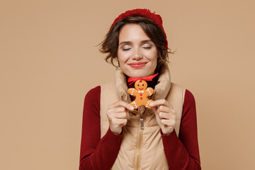 Young satisfied smiling caucasian woman 20s wears red turtleneck vest beret hold in hand sniff ginger cookie biscuit isolated on plain pastel beige background studio portrait People lifestyle concept