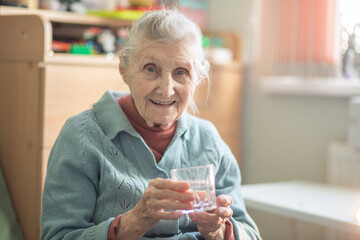 An elderly woman holds a glass glass with water in her hand, health care, grandmother drinks water for health, retirees at home during quarantine.
 - obrazy, fototapety, plakaty