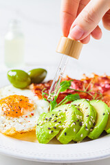 Cannabis essential oil CBD drip into food. Keto breakfast concept. Fried egg with avocado and fried bacon in white plate.