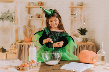 cute little girl in a green Halloween costume of a witch or fairy prepares a pumpkin pie, barm brack in a festive decorated kitchen. space for text. High quality photo