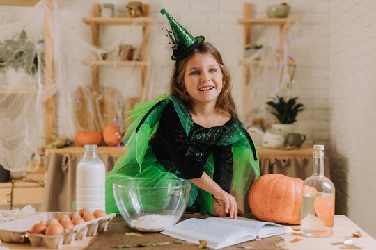 cute little girl in a green Halloween costume of a witch or fairy prepares a pumpkin pie, barm brack in a festive decorated kitchen. space for text. High quality photo