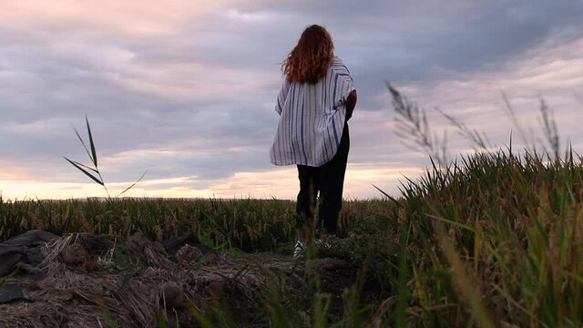 Shirtless woman admiring and reflecting on a landscape of a rice field as the breeze gives her.
