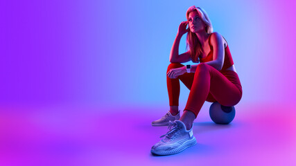 Woman resting after cross training at gym on fitness ball. Modern neon light background. Woman taking break from physical training