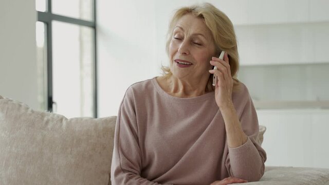 Smiling elderly woman talking on a mobile phone at home, relaxing on the sofa in the living room