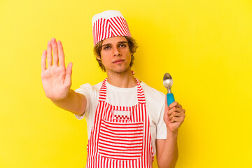 Young ice cream maker man with makeup holding spoon isolated on yellow background  standing with...