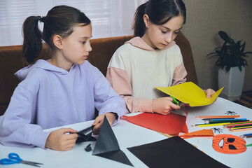 Children in a dim room are sitting at a table, cutting out a garland of orange and black paper for Halloween.
