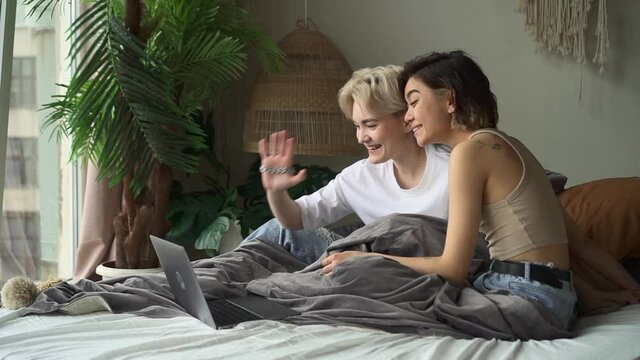 Lesbian couple at videocall. Spbd Blonde young woman and Asian wife greet friends waving hands at online chat via laptop sitting on large bed at home