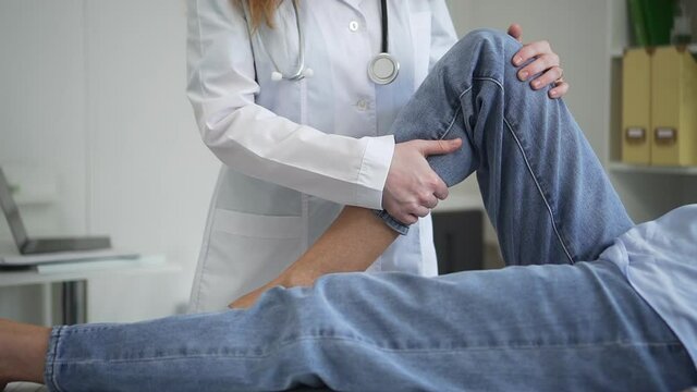 Doctor conducts appointment with patient. Spbd Skilled lady physiatrist in white robe bends leg doing rehabilitation exercises to woman in hospital room closeup