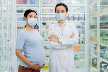 pharmacist in medical mask standing with crossed arms near asian pregnant woman in drugstore