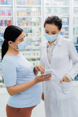 Fototapeta na wymiar asian pregnant woman in medical mask pointing at smartphone near pharmacist in drugstore