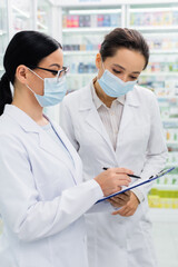 asian pharmacist in glasses and medical mask writing on clipboard near colleague