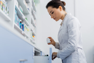 tattooed pharmacist in white coat holding bottle with medication while opening drawer in drugstore