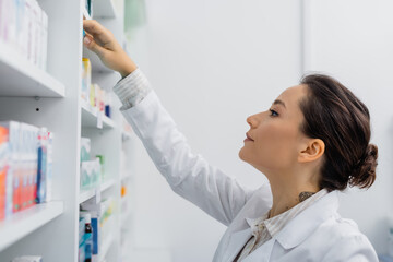 tattooed apothecary in white coat reaching shelf with medication in drugstore