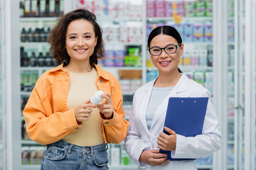 asian pharmacist in glasses holding clipboard near cheerful customer with bottle in drugstore