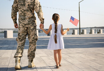 Soldier and his little daughter with American flag outdoors, back view. Veterans Day in USA