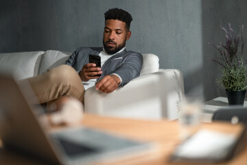 Fototapeta na wymiar Young african american businessman with smartphone sitting on sofa indoors in office