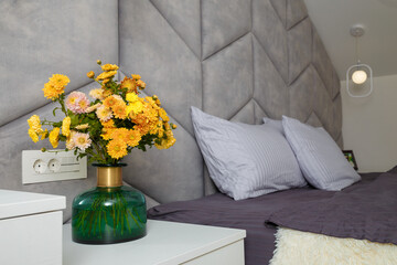 Beautiful grey bedroom, bed, pillows, soft fabric headboard, autumn bouquet of yellow chrysanthemums in green vase near. Modern interior design