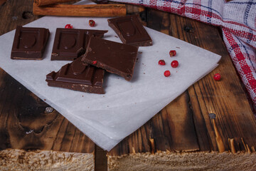 Broken bar of dark bitter chocolate with chilly pepper, cinnamon, red pea pepper inside on paper, wooden board background. Unusual taste