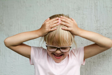 The child shows that he has a headache. A little girl with blonde hair in a pink T-shirt holding her head with her hands and showing emotions of pain