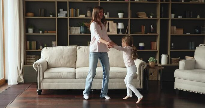 Having fun with mum. Candid energetic young mom nanny dancing at living room with junior school age girl daughter. Happy friendly sisters adult elder kid younger hold hands jump laugh at home go crazy
