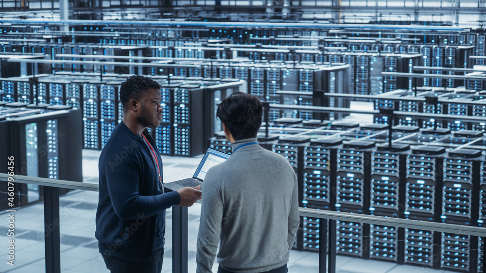 Poster Two Multiethnic Male Data Center System Administrators and IT Specialists Talk, Use Laptop. Information Technology Engineers work on Cyber Security Network Protection in Cloud Computing Server Farm.