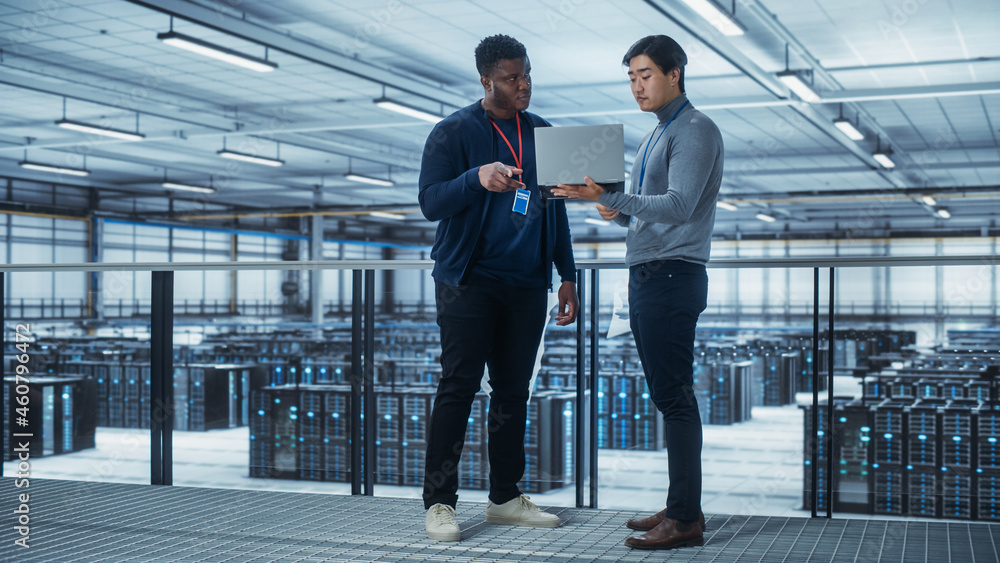 Poster Two Multiethnic Male Data Center System Administrators and IT Specialists Talk, Use Laptop. Information Technology Engineers work on Cyber Security Network Protection in Cloud Computing Server Farm.
