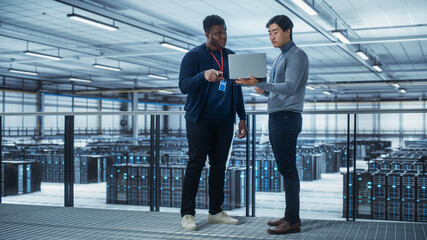 Two Multiethnic Male Data Center System Administrators and IT Specialists Talk, Use Laptop. Information Technology Engineers work on Cyber Security Network Protection in Cloud Computing Server Farm.