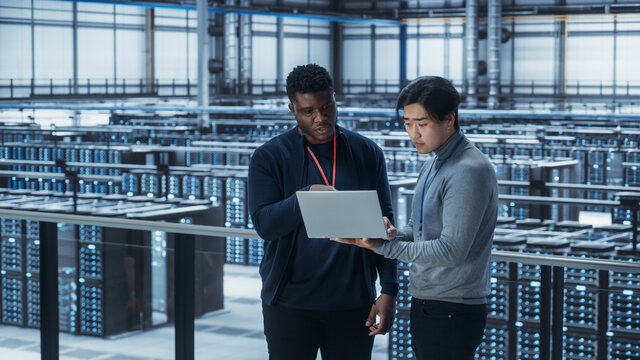 Two Multiethnic Male Data Center System Administrators and IT Specialists Talk, Use Laptop. Information Technology Engineers work on Cyber Security Network Protection in Cloud Computing Server Farm.