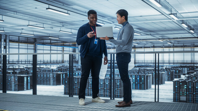 Two Multiethnic Male Data Center System Administrators and IT Specialists Talk, Use Laptop. Information Technology Engineers work on Cyber Security Network Protection in Cloud Computing Server Farm.