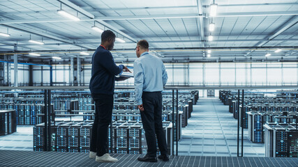 Male Data Center System Administrators and African American IT Specialists Talk, Use Laptop. Information Technology Engineers work on Cyber Security Network Protection in Cloud Computing Server Farm.