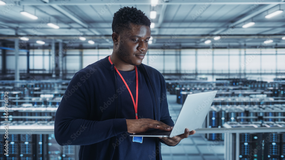 Poster Portrait of a Data Center Engineer Using Laptop Computer. Server Room Specialist Facility with African American Male System Administrator Working with Data Protection Network for Cyber Security.