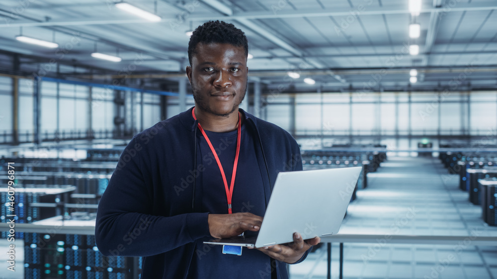 Poster Portrait of a Data Center Engineer Using Laptop Computer. Server Room Specialist Facility with African American Male System Administrator Working with Data Protection Network for Cyber Security.