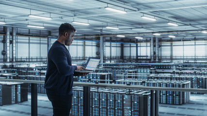 Data Center Engineer Using Laptop Computer. Server Farm Cloud Computing Specialist Facility with African American Male System Administrator Working with Data Protection Network for Cyber Security.