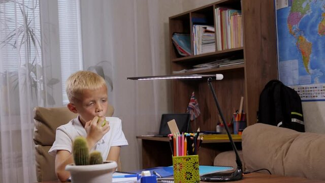 A little boy in a white T-shirt is sitting at his desk, doing homework and eating a green apple. In the background there are bookshelves and a home interior.