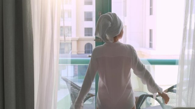 Woman with a towel on the head after a shower opens the curtains at the hotel window early in the morning. Sun is shining brightly outside. A vacation begining with good weather and a great emotions.